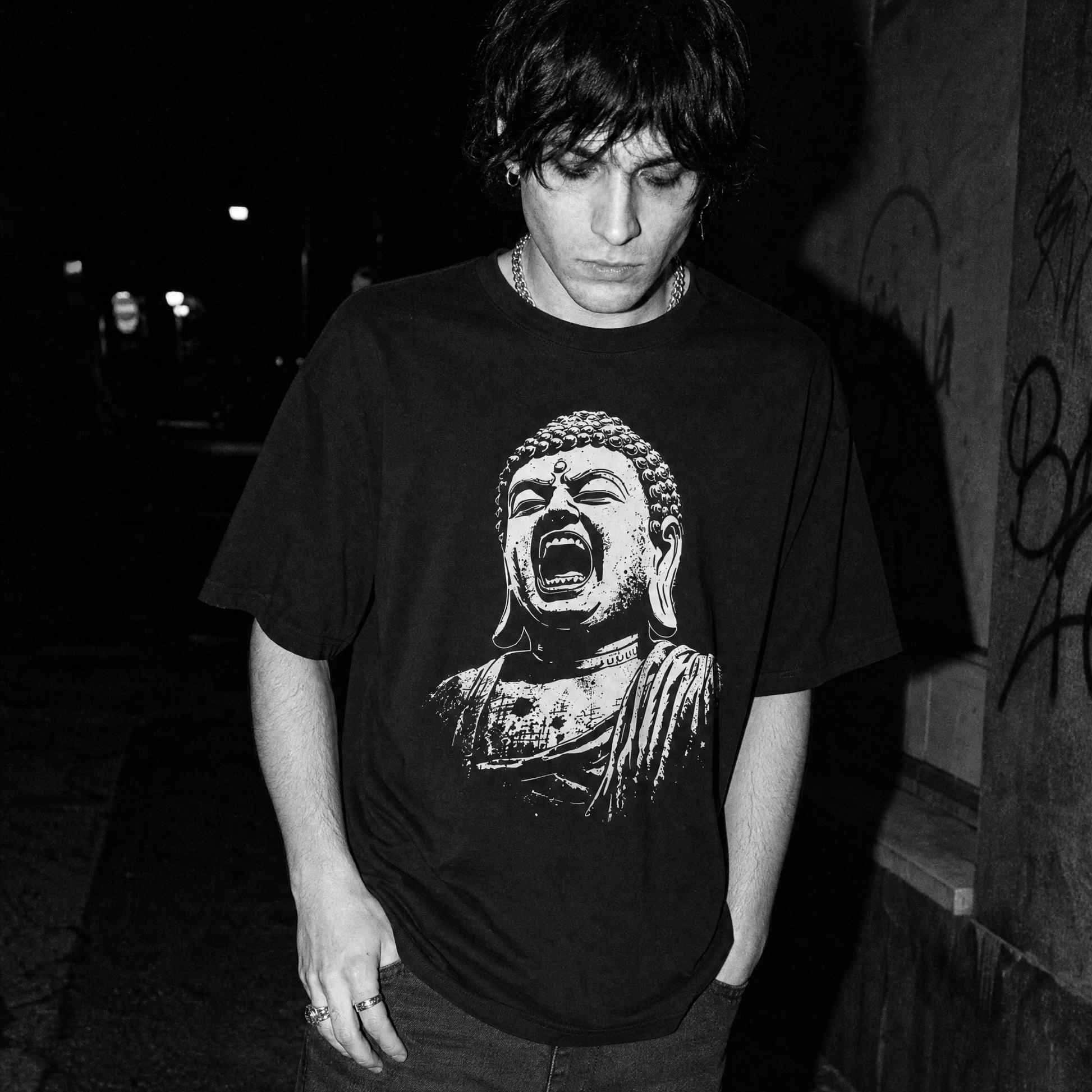 A male wearing a black short-sleeve graphic T-shirt with a large white screaming Buddha bust print on the front, photographed against a dark brick and metal gate background.