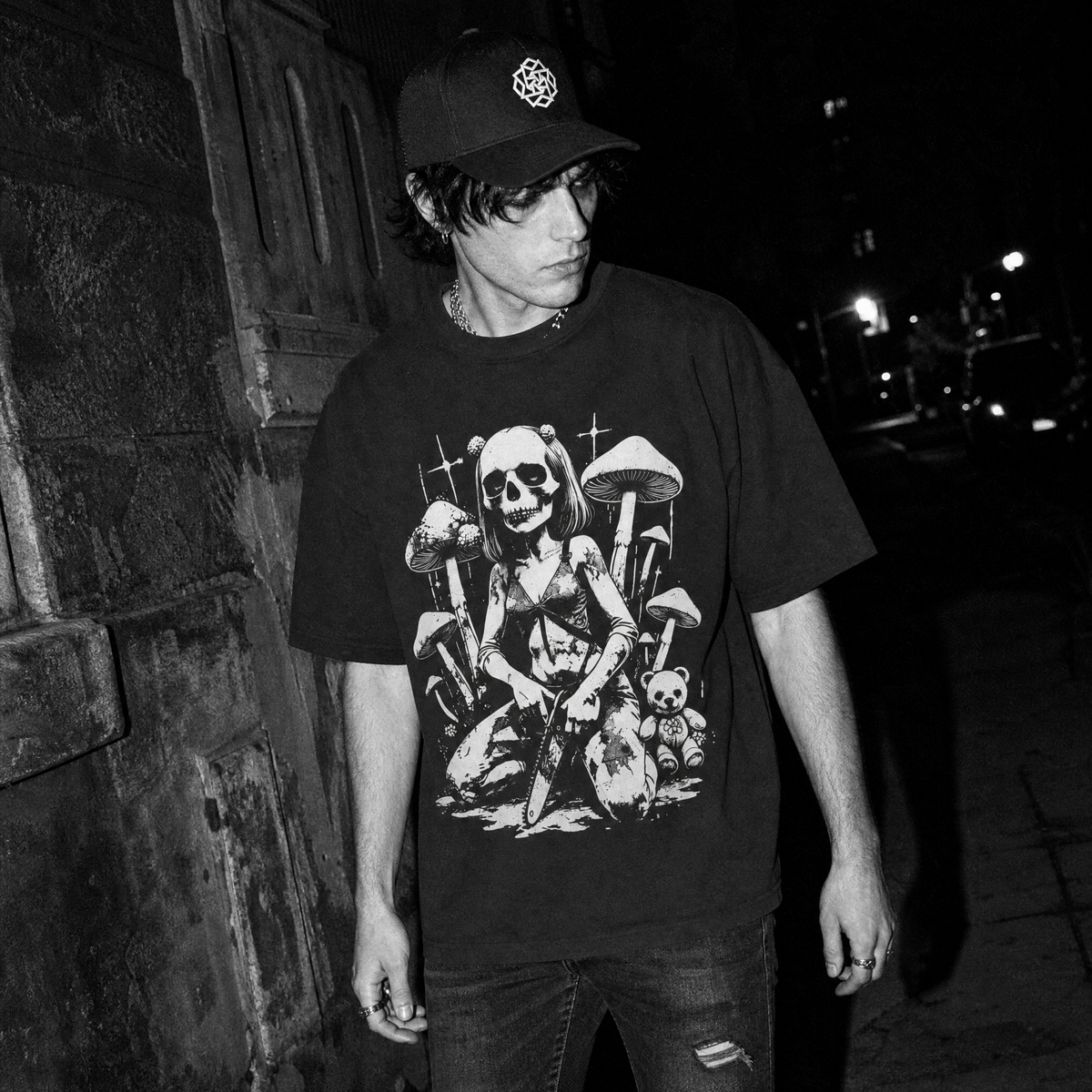 A person standing outdoors at night wearing a black graphic T-shirt with a  print of a skull-headed girl gripping a chainsaw surrounded by mushrooms and a teddy bear, a dark baseball cap, and jeans.