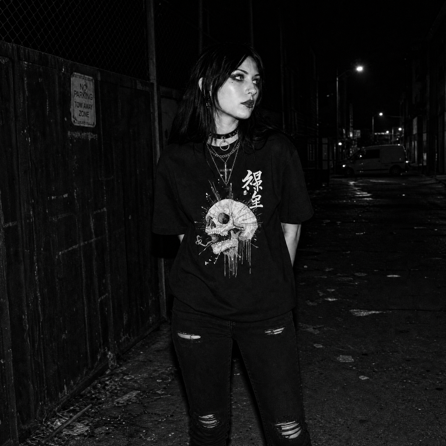 A woman standing at night on a dark street beside a wooden fence, wearing a black graphic T-shirt with a white skull print and Kanji characters, along with distressed black jeans.