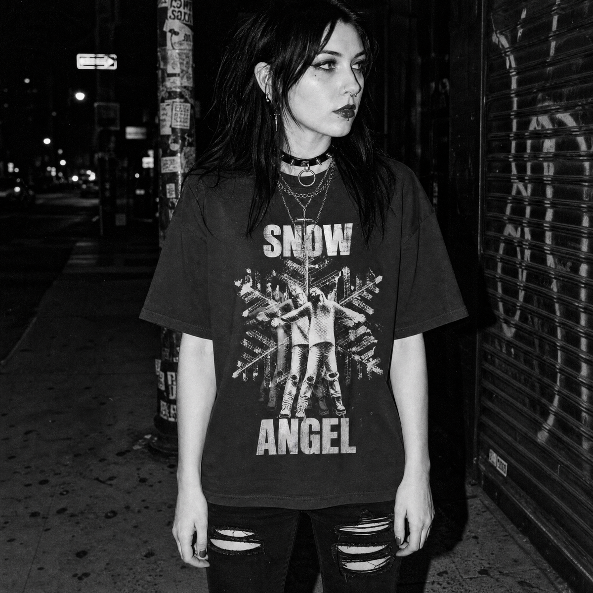 A woman standing on a city sidewalk at night wearing an oversized black graphic T-shirt with the words 'SNOW ANGEL' and a distressed figure print, paired with ripped black jeans and layered necklaces.