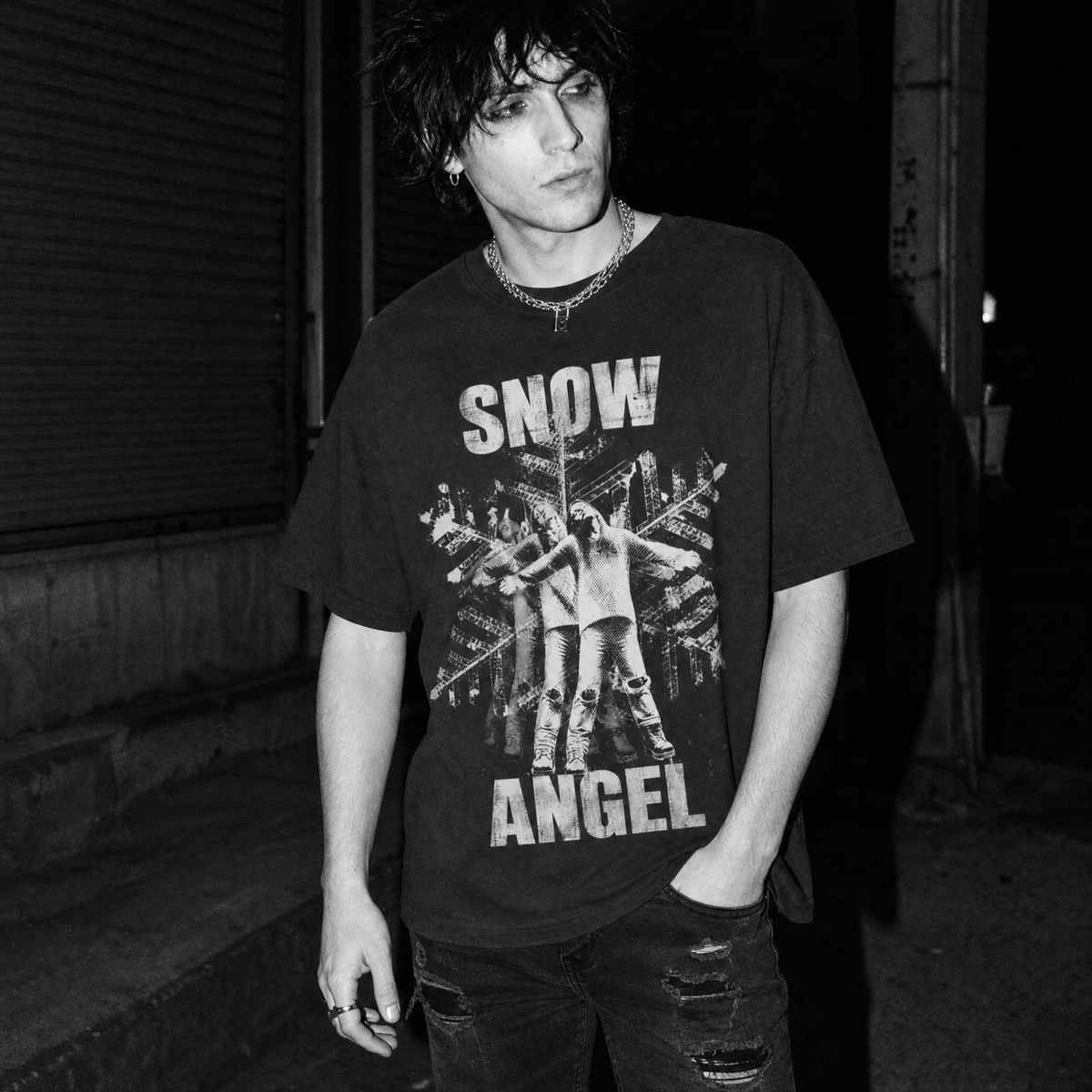 A man wearing a black baseball cap and a black graphic t-shirt with 'SNOW ANGEL' text and a snowflake design, standing outdoors at night against a metal gate.