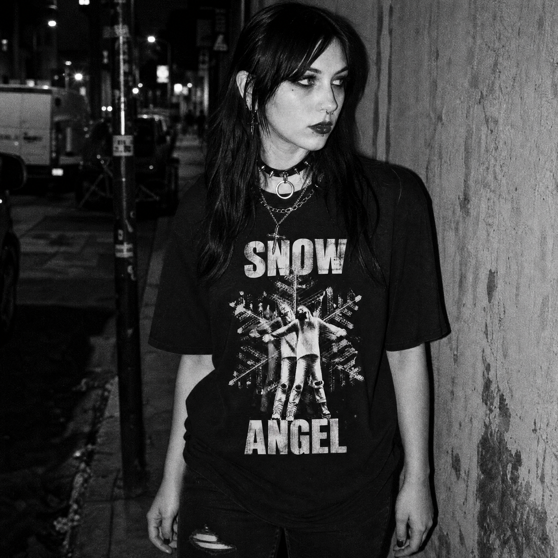 A woman posing on a city sidewalk at night wearing a black graphic t-shirt with 'SNOW ANGEL' text and a snowflake design, paired with distressed black jeans.