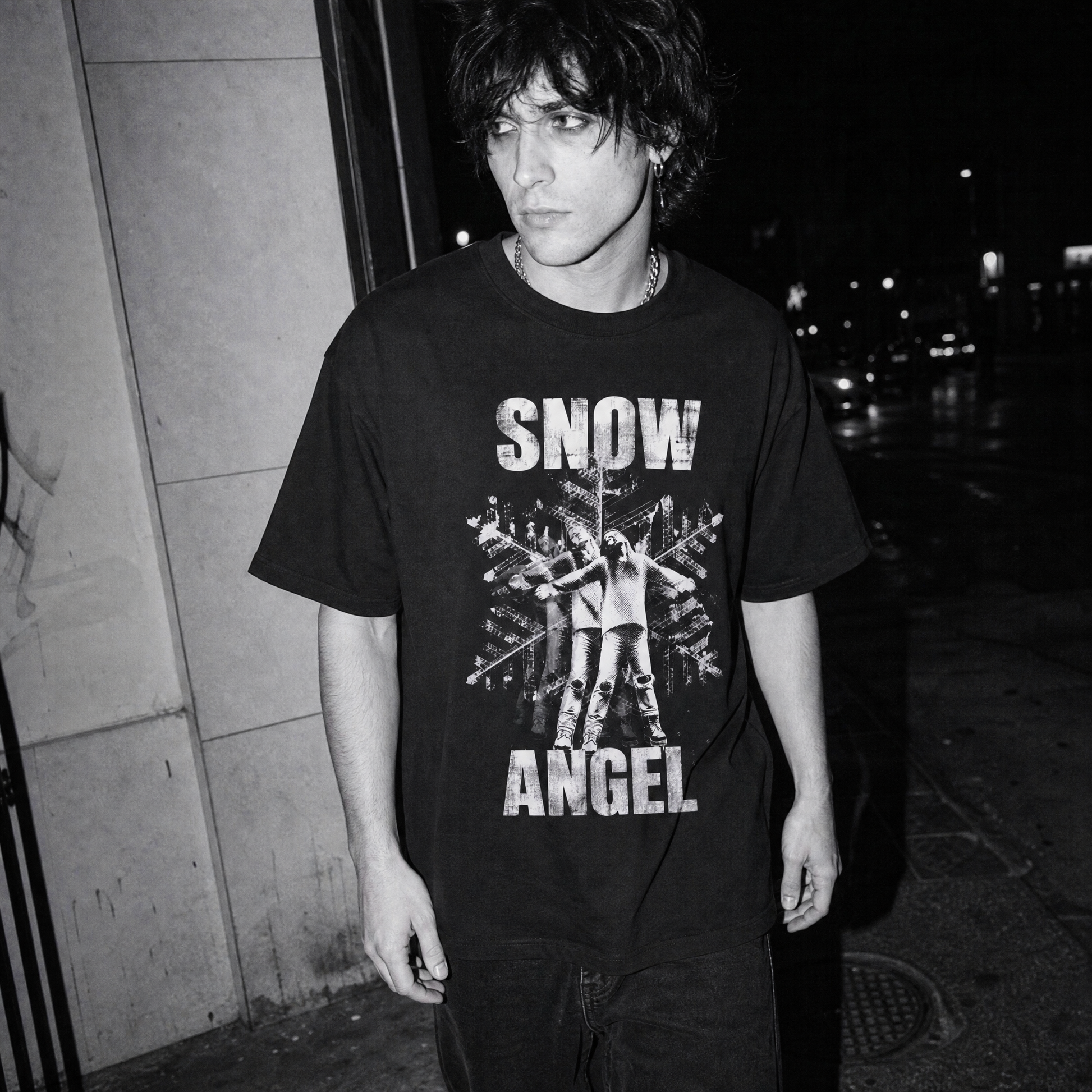 A black and white street-style photograph of a man wearing a graphic t-shirt  of a woman with arms spread out on top of a giant snowflake with the words 'SNOW ANGEL', standing outdoors at night.