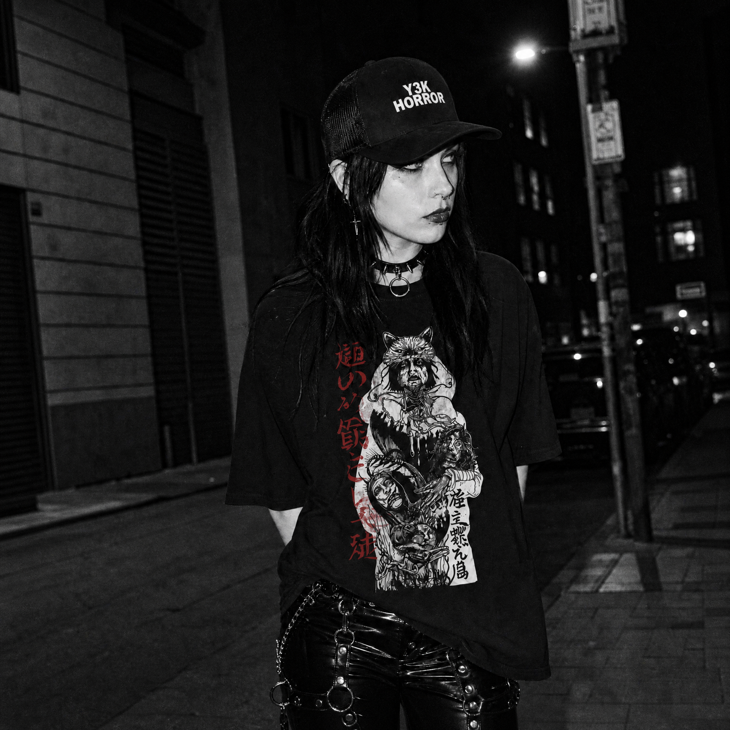 A woman standing on a city street at night wearing a black graphic T-shirt with a large illustration of a warrior carrying three heads, red vertical Kanji text, a black cap with white text reading 'Y3K Horror', and shiny black bondage pants.