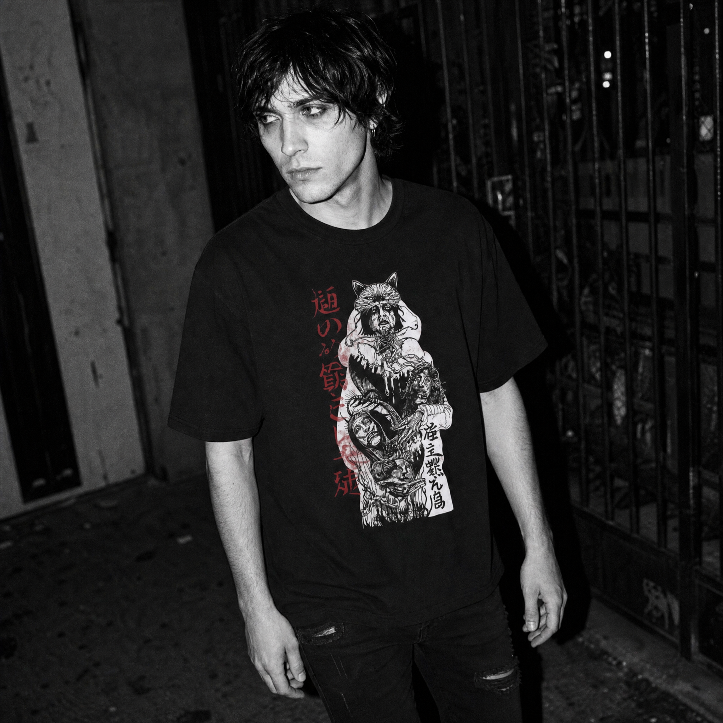 A man wearing a black graphic t-shirt with a detailed red, white, and gray print of a warrior with a wolf head carrying three heads and Kanji text, standing in a dark alleyway at night.
