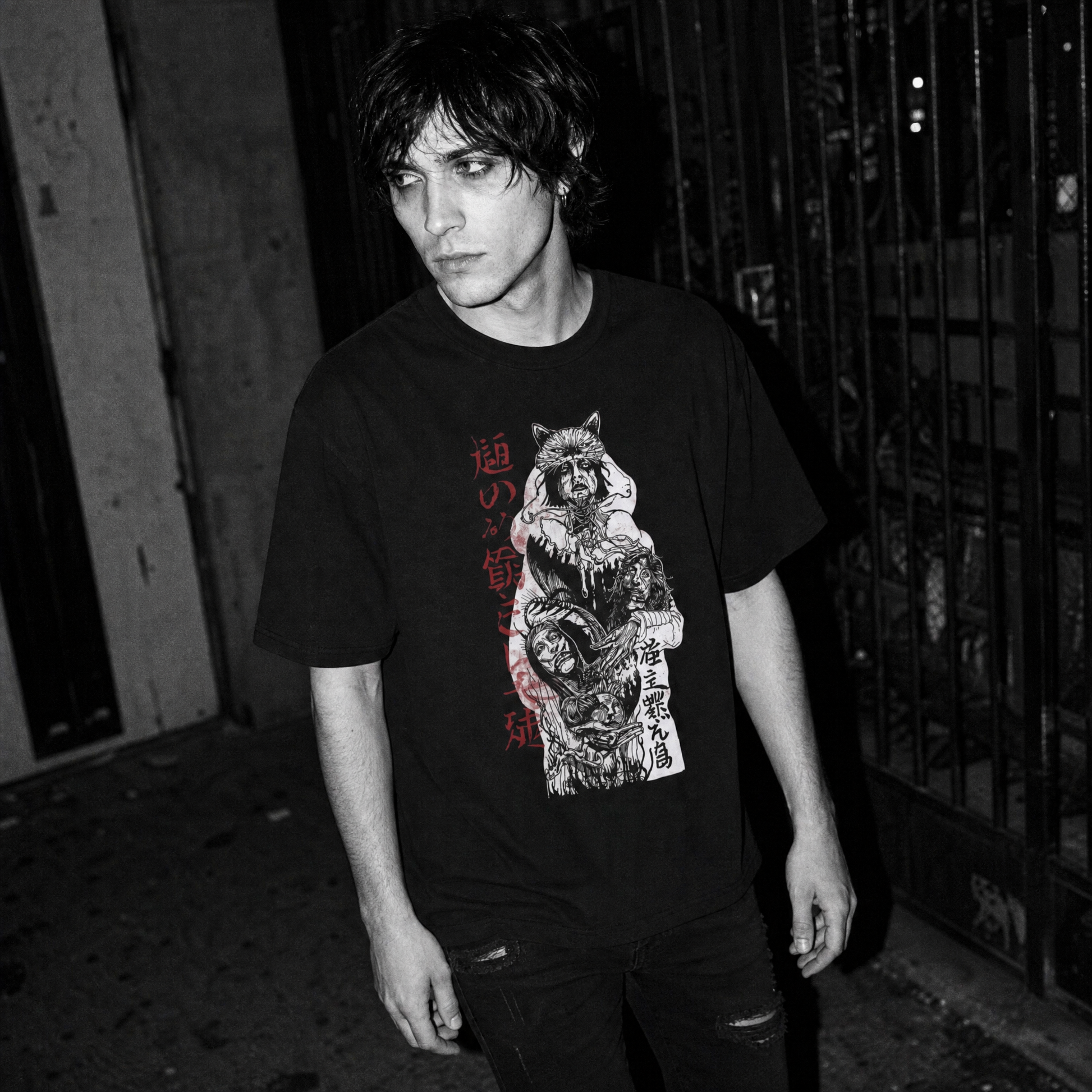 A man wearing a black graphic t-shirt with a detailed red, white, and gray print of a warrior with a wolf head carrying three heads and Kanji text, standing in a dark alleyway at night.