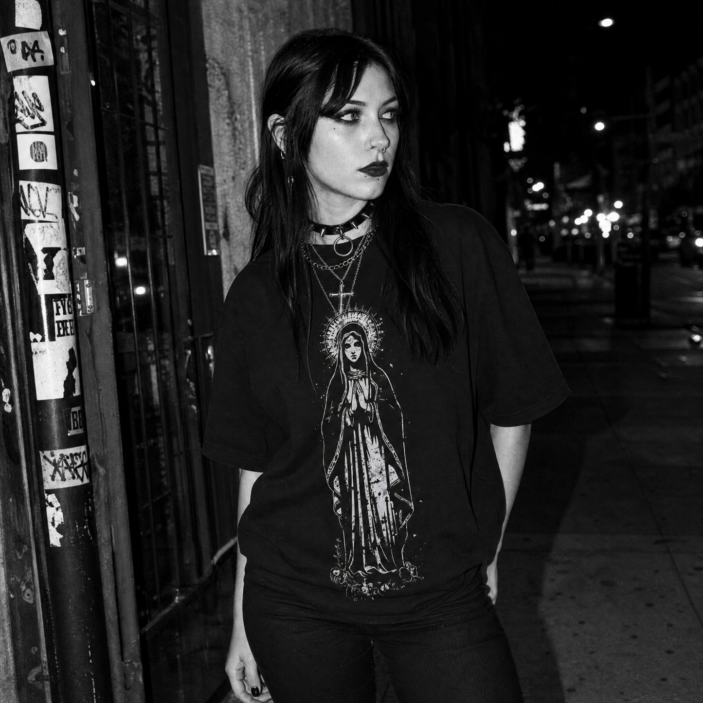 A woman stands on a city sidewalk at night wearing an oversized black graphic T-shirt with a detailed white religious print of the Virgin Mary on the front, paired with dark pants and layered necklaces.