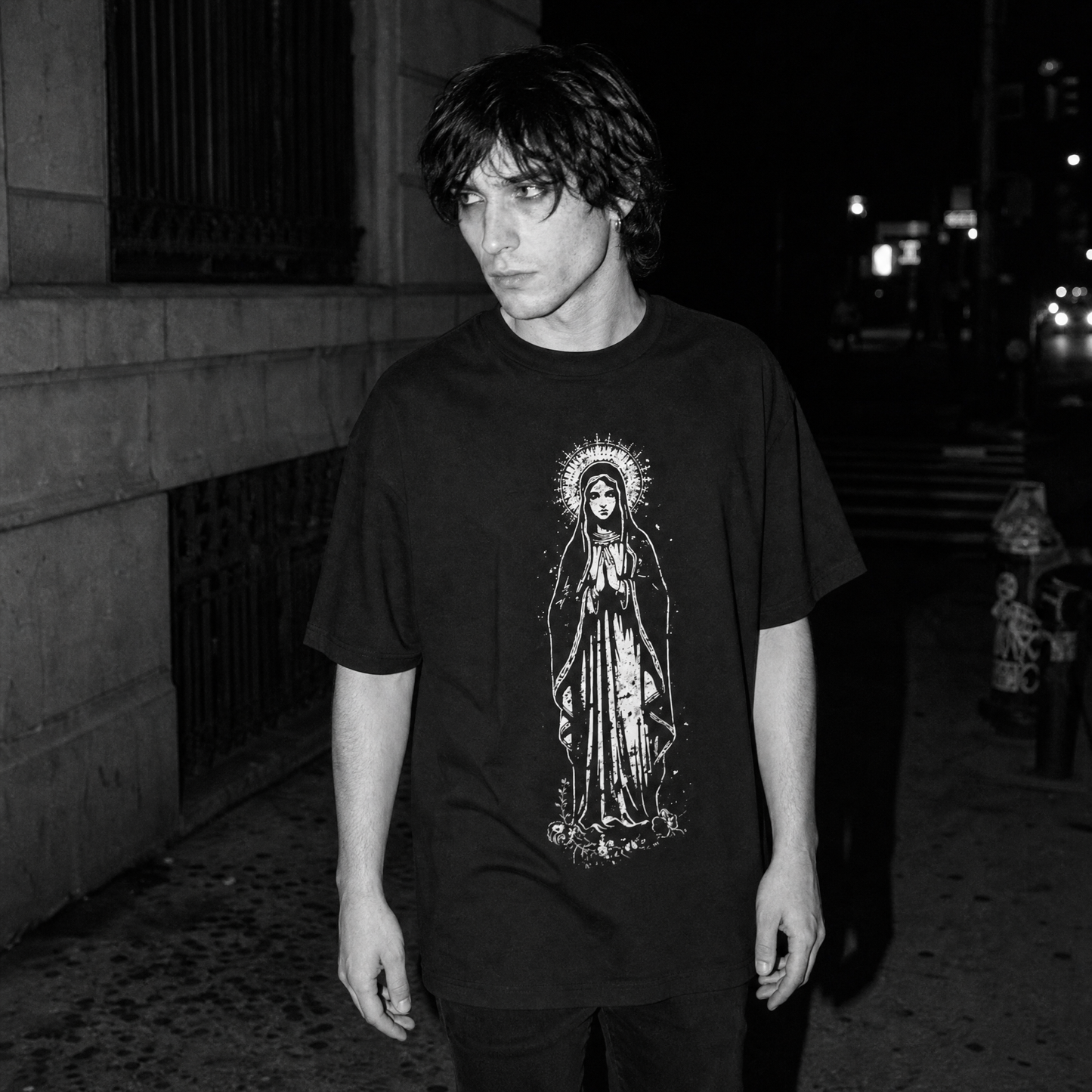 A man wearing a black oversized graphic t-shirt with a white, distressed line art print of the Virgin Mary, standing on a dark city street at night.