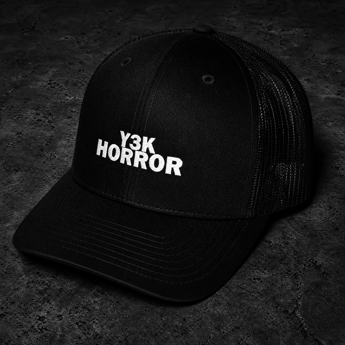 A black trucker-style cap with white embroidered text reading 'Y3K HORROR' on the front, photographed on a dark textured surface.