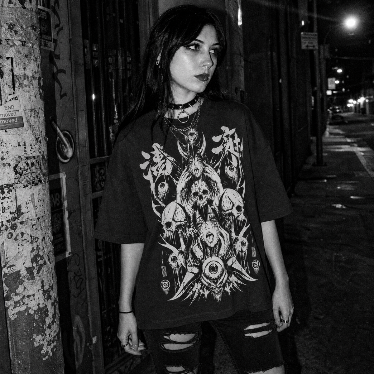 A woman wearing an oversized black graphic t-shirt with a detailed white gothic design featuring skulls, occult symbols, and a central eye motif, paired with ripped black jeans, standing on a city street at night.