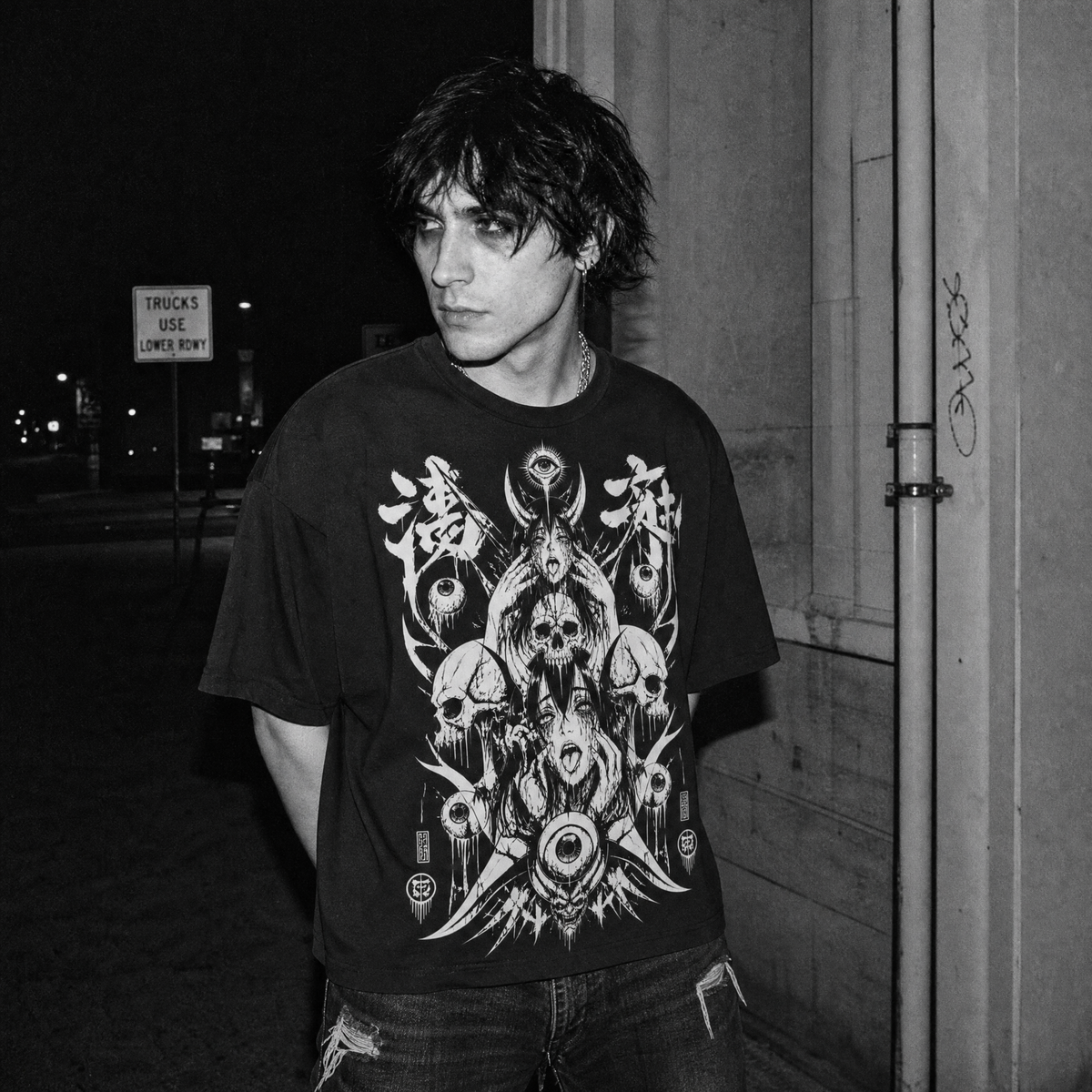 A man wearing a black graphic t-shirt with a detailed white and gray design featuring skulls, eyes, and occult symbols, standing outdoors at night.