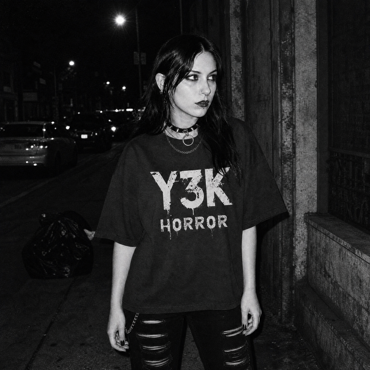 Black-and-white nighttime street photo of a female model wearing a black oversized T-shirt with distressed white “Y3K HORROR” text on the front.